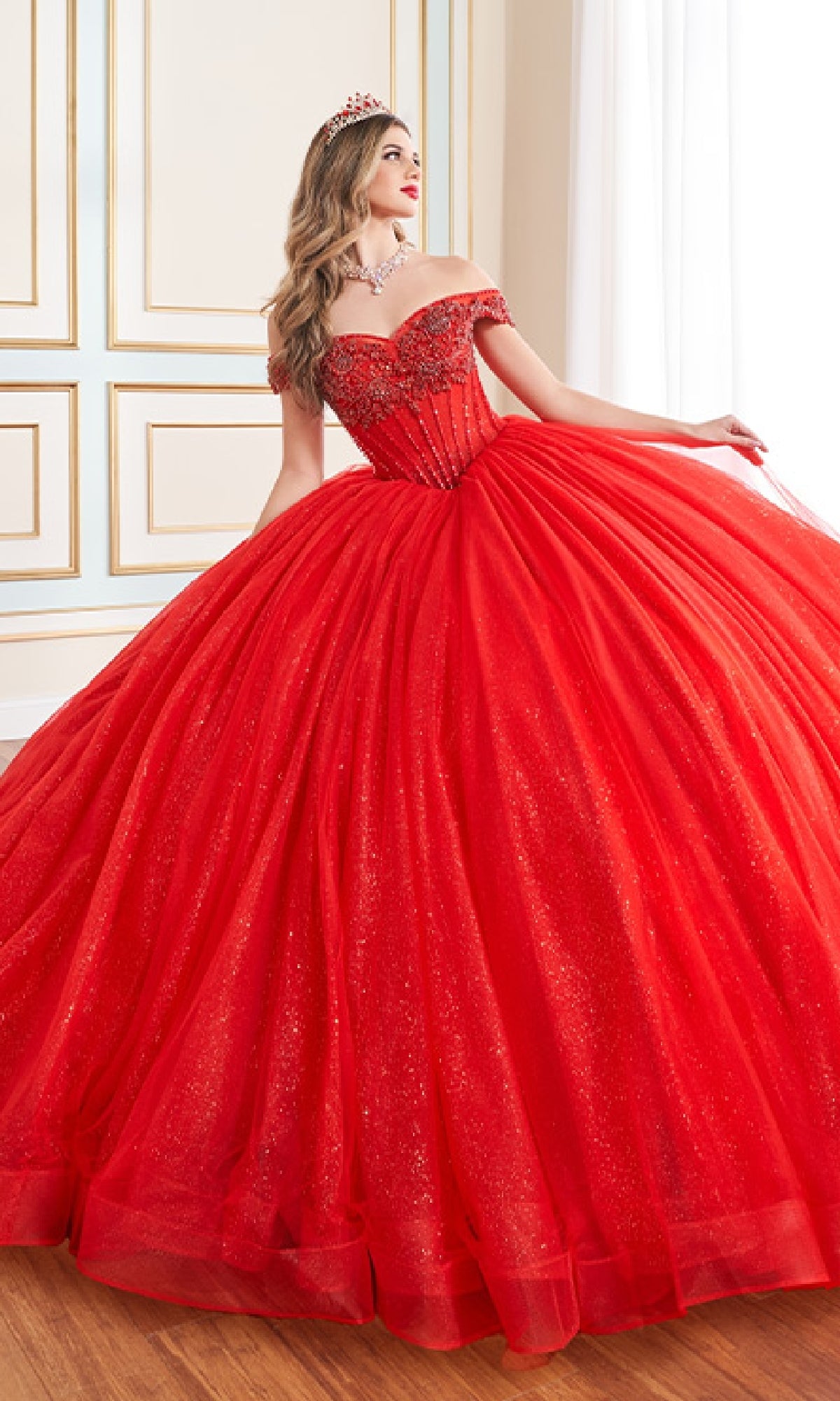 Wearing the Quinceanera Dress PR30180 By Princesa, a person in a sparkling red glitter tulle ball gown with an off-the-shoulder neckline and tiara stands indoors near a window, gazing to the side.